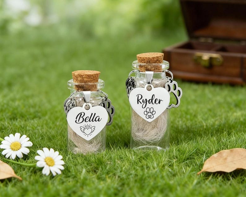 May include: Two small glass bottles with cork stoppers, filled with a light-coloured material. Each bottle has a heart-shaped tag with a name and paw print. One says "Bella" and the other says "Ryder".