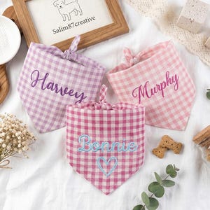 May include: Three triangular pet bandanas in gingham patterns. One is light purple with the name Harvey in dark purple embroidery. One is light pink with the name Murphy in pink embroidery. One is dark pink with Bonnie and a heart in light blue.