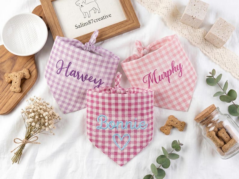 May include: Three triangular pet bandanas in gingham patterns. One is light purple with the name Harvey in dark purple embroidery. One is light pink with the name Murphy in pink embroidery. One is dark pink with Bonnie and a heart in light blue.