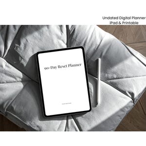 May include: A digital tablet with a black bezel displays the text "90-Day Reset Planner" in a minimalist font. A white stylus rests beside the tablet on a gray, textured surface. The image also includes the text "Undated Digital Planner iPad & Printable".