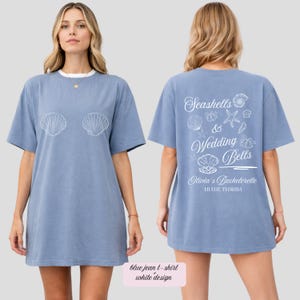 以下が含まれることがあります： 前面に白い貝殻のデザイン、背面に「Seashells & Wedding Bells, Olivia's Bachelorette, Miami, Florida」と書かれた、青いジーンズのTシャツ。 シャツは白い襟が付いています。