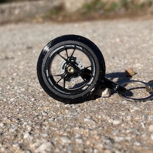 Puede incluir: Un llavero de rueda de motocicleta negra con un llavero plateado y una correa trenzada negra. La rueda tiene un diseño detallado con radios y un disco de freno plateado. El llavero está sobre una superficie gris texturizada.
