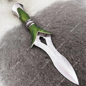 Op de afbeelding: Een decoratieve dolk met een gepolijst zilveren lemmet en een groene handgreep. De handgreep heeft een zilveren bolster en een decoratieve zilveren band. De dolk wordt getoond op een grijs en wit vachtachtig oppervlak.