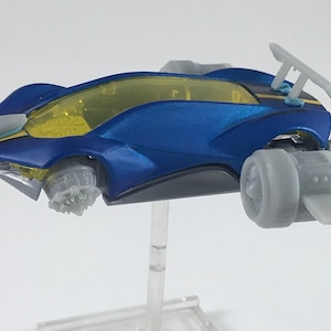 May include: A blue toy car with a yellow windshield and gray wheels. The car has a spoiler and a streamlined design. The toy car is on a clear stand.
