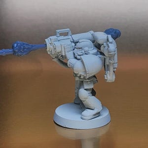 May include: A light gray miniature figure of a soldier in armor, holding a large weapon with a blue energy blast. The figure is standing on a circular base. The weapon has intricate details.