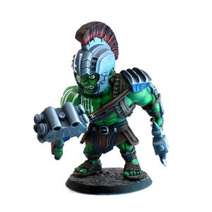 Puede incluir: Figura verde de un personaje musculoso con casco y armadura. La figura sostiene un arma grande y gris en cada mano. La figura tiene una expresión decidida y está de pie sobre una base negra.