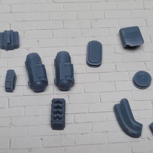 May include: An assortment of small, gray plastic model parts, including what appear to be engine components, exhaust pipes, and other mechanical elements. The parts are arranged on a white brick surface.