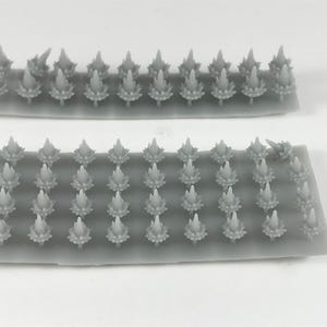 May include: Two gray resin sprue sets, each holding multiple small, stylized, floral-like components. The components have a textured, layered appearance with pointed tips. The sets are arranged on a white surface, suitable for miniature crafting or model making.