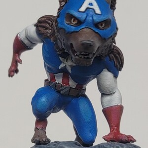 Puede incluir: Figura de una criatura parecida a un lobo con un disfraz de Capitán América, arrodillada sobre una roca gris. El disfraz es azul con una estrella blanca, rayas rojas y blancas y una máscara azul con una "A" blanca. La figura tiene pelaje marrón y guantes y botas rojas.