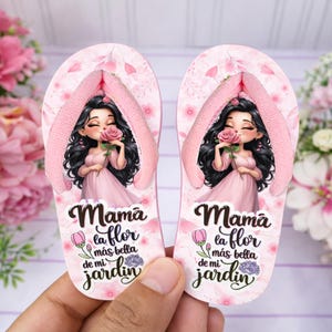 Può includere: Infradito rosa con un design floreale e la scritta "Mama la flor más bella de mi jardín." Ogni infradito presenta una donna dei cartoni animati che tiene una rosa. I cinturini sono rosa.