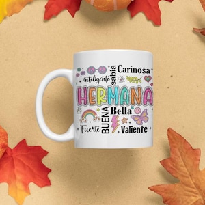Peut inclure: Mug en céramique blanche avec le mot "HERMANA" en lettres colorées. Le mug comporte du texte et des graphiques supplémentaires, dont un arc-en-ciel, un papillon et des étoiles. Le mug est un cadeau pour une sœur.
