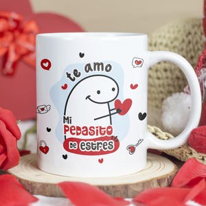 Puede incluir: Taza de cerámica blanca con un personaje de dibujos animados sosteniendo un corazón rojo, rodeado de corazones y el texto "te amo Mi Pedacito de estres". La taza está sobre un posavasos de madera, con una rosa roja y pétalos.