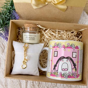Puede incluir: Una caja de regalo con un lazo dorado contiene una taza, una vela y un llavero. La taza amarilla presenta un personaje de dibujos animados con pelo largo negro y un fondo rosa. La vela está en un pequeño tarro de cristal con tapa de corcho. El llavero tiene una letra "S" dorada.