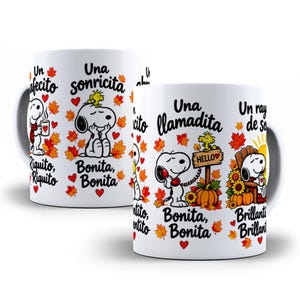Può includere: Una tazza in ceramica bianca con illustrazioni di Snoopy e Woodstock circondate da foglie autunnali, cuori rossi e testi in spagnolo come Un cafecito, Una sonricita, Una llamadita e Un rayito de sol, con zucche e girasoli.