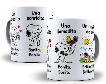 Puede incluir: Una taza de cerámica blanca con ilustraciones de Snoopy y texto en español. Las escenas muestran a Snoopy con una taza de café, sonriendo, hablando por teléfono con Woodstock y sentado junto a un sol. El texto incluye Un cafecito, Una sonricita, Una llamadita y Un rayito de sol.