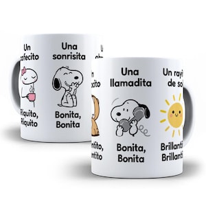 May include: A white ceramic mug featuring cartoon illustrations of Snoopy and other characters, accompanied by Spanish text including phrases like Un cafecito, Una sonrisita, Una llamadita, and Un rayito de sol, with colourful accents like a pink cup and yellow sun.