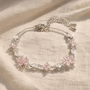 Peut inclure: Un bracelet argenté orné d'un motif floral avec des cristaux roses et transparents. Il est doté d'une chaîne délicate et d'un fermoir coulissant réglable. Les fleurs sont composées de cristaux en forme de marquise et ronds.