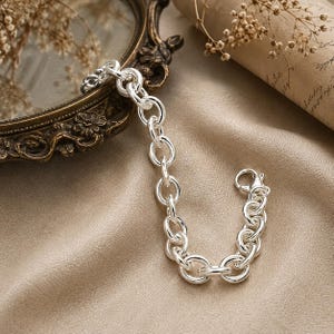 Può includere: Un bracciale a catena in argento lucido caratterizzato da una serie di maglie circolari uniformi e arrotondate collegate tra loro. Il bracciale include una chiusura a moschettone sicura a un'estremità, con una finitura metallica brillante e riflettente.
