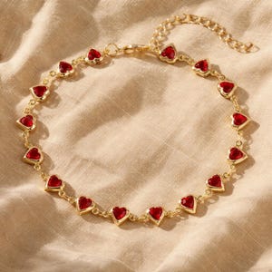 May include: Gold-colored anklet featuring heart-shaped red gemstones. The anklet has a delicate chain design with multiple heart-shaped stones evenly spaced around the circumference. The anklet is displayed on a beige fabric background.