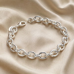 May include: A polished silver chain link bracelet featuring thick, rounded oval links connected in a continuous loop. The bracelet includes a secure lobster claw clasp and a small circular tag attached near the closure, showcasing a bright metallic finish.