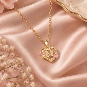 Könnte beinhalten: Eine goldfarbene Halskette mit einem herzförmigen Anhänger, der in der Mitte den Buchstaben M trägt. Der Herzrahmen ist an den Seiten mit kleinen rosa Blumenakzenten verziert. Der Anhänger hängt an einer feinen goldfarbenen Gliederkette.