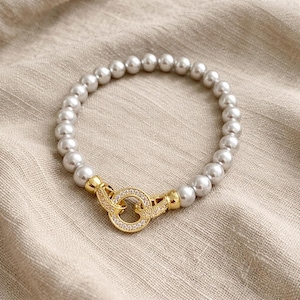May include: A pearl bracelet with a gold clasp. The bracelet features light grey pearls strung together. The clasp is gold with a circular design and small, clear stones. The bracelet is displayed on a neutral-coloured fabric.