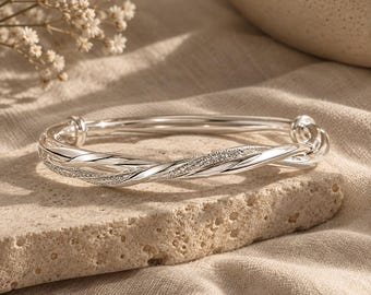 Könnte beinhalten: Ein silberfarbener Armreif mit gedrehtem Design, bestehend aus abwechselnd glatten, polierten Abschnitten und texturierten, glitzernden Segmenten. Der Armreif hat eine kreisförmige Form mit abgerundeten Enden, die sich an der Vorderseite treffen.