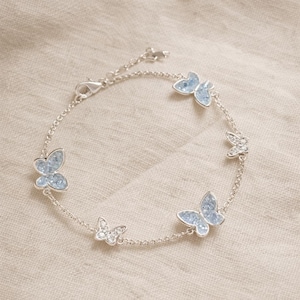 Pode incluir: Pulseira de prata com seis pingentes de borboleta. Cada borboleta é adornada com cristais azuis claros. A pulseira é conectada por uma corrente fina e tem um fecho. Comprimento aproximado: 18 cm.