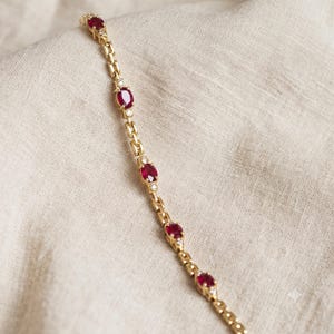 Puede incluir: Una pulsera de cadena dorada que presenta una serie de rubíes de color rojo intenso con corte ovalado, cada uno flanqueado por pequeñas piedras de acento transparentes de corte redondo. La pulsera tiene eslabones dorados pulidos en línea.