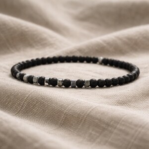 May include: A black beaded bracelet with silver-colored metal accents. The bracelet features small, round black beads interspersed with cylindrical and square metal beads. The bracelet is displayed on a neutral-toned fabric background.