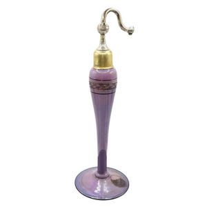 Può includere: Un flacone di profumo decorativo vintage con un corpo in vetro color lavanda e una parte superiore in metallo argentato. Il flacone presenta un beccuccio curvo e una fascia color oro vicino alla parte superiore, con un design ornato. La base è di colore lavanda abbinato.