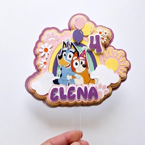Peut inclure: Décoration de gâteau multicouche avec les personnages de Bluey et Bingo se câlinant devant un arc-en-ciel. La décoration comprend le chiffre 4, des ballons, des marguerites, un soleil, des nuages et le nom "ELENA".