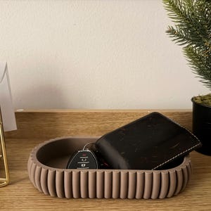 May include: A brown, ribbed, oval-shaped tray holds a black wallet and a set of keys. A small potted plant and a brown leather wallet holder are also visible on a wooden surface.