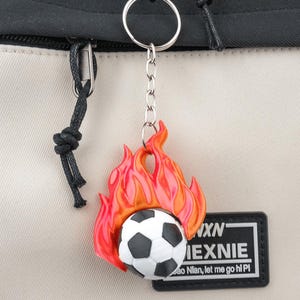 May include: A keychain featuring a soccer ball engulfed in flames, with a black and white design. The keychain is attached to a silver keyring and chain. A black and white patch with text is visible.
