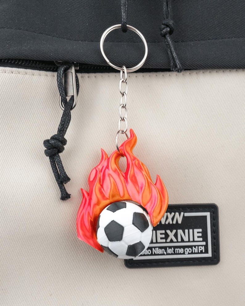 May include: A keychain featuring a soccer ball engulfed in flames, with a black and white design. The keychain is attached to a silver keyring and chain. A black and white patch with text is visible.