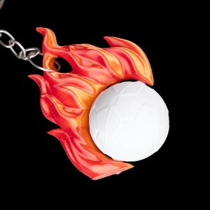 May include: A keychain featuring a white soccer ball design with a red and orange flame accent. The keychain is attached to a silver chain against a black background.