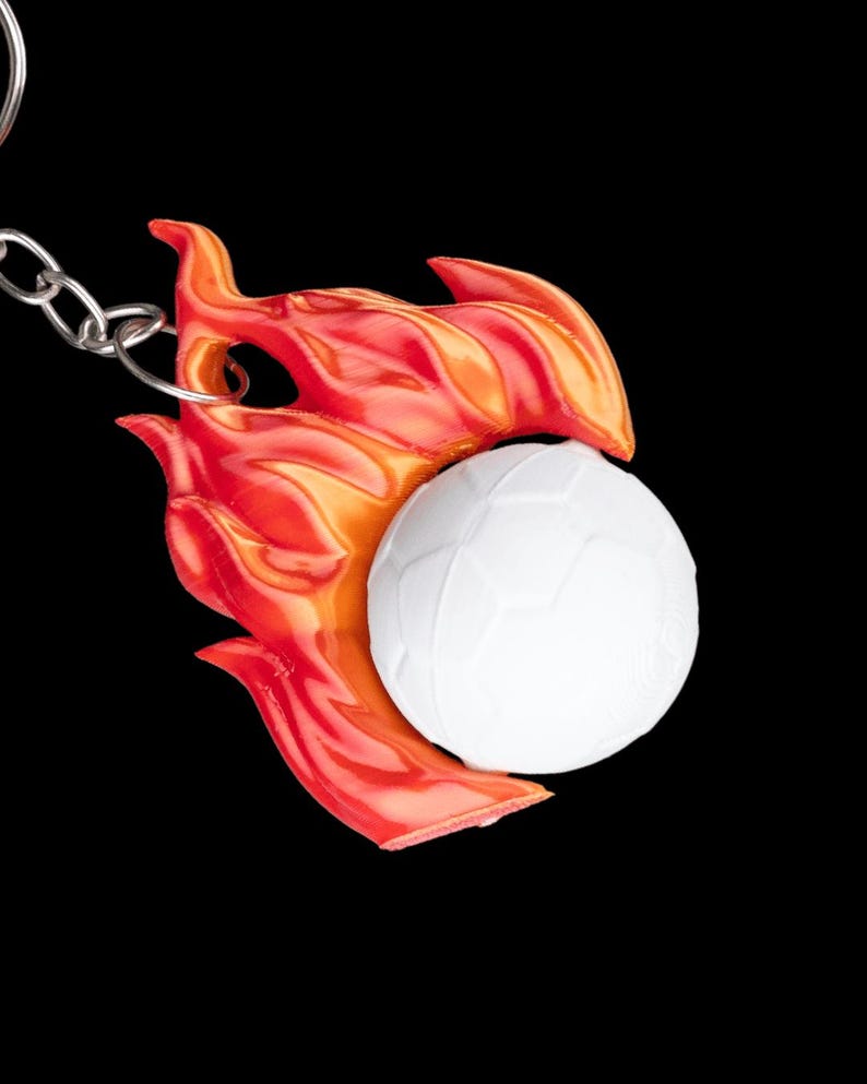 May include: A keychain featuring a white soccer ball design with a red and orange flame accent. The keychain is attached to a silver chain against a black background.