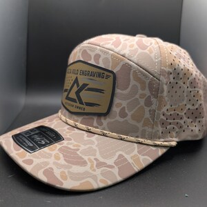 May include: A tan and beige camouflage patterned baseball cap with a perforated back panel. The front features a rectangular patch with the text "OLD KILO ENGRAVING" and "VETERAN OWNED" alongside a logo. A decorative rope trim sits above the brim.