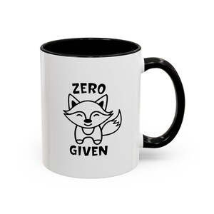 May include: A white ceramic mug with a black interior and a black handle. The front of the mug features a black line art illustration of a small fox with closed eyes and the text ZERO GIVEN printed in a bold, sans-serif font above and below it.