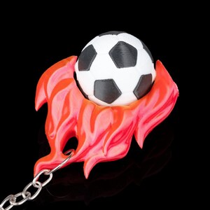 May include: A soccer ball keychain with a black and white soccer ball design, set within a red and orange flame-like shape. The keychain has a silver chain and a black background.