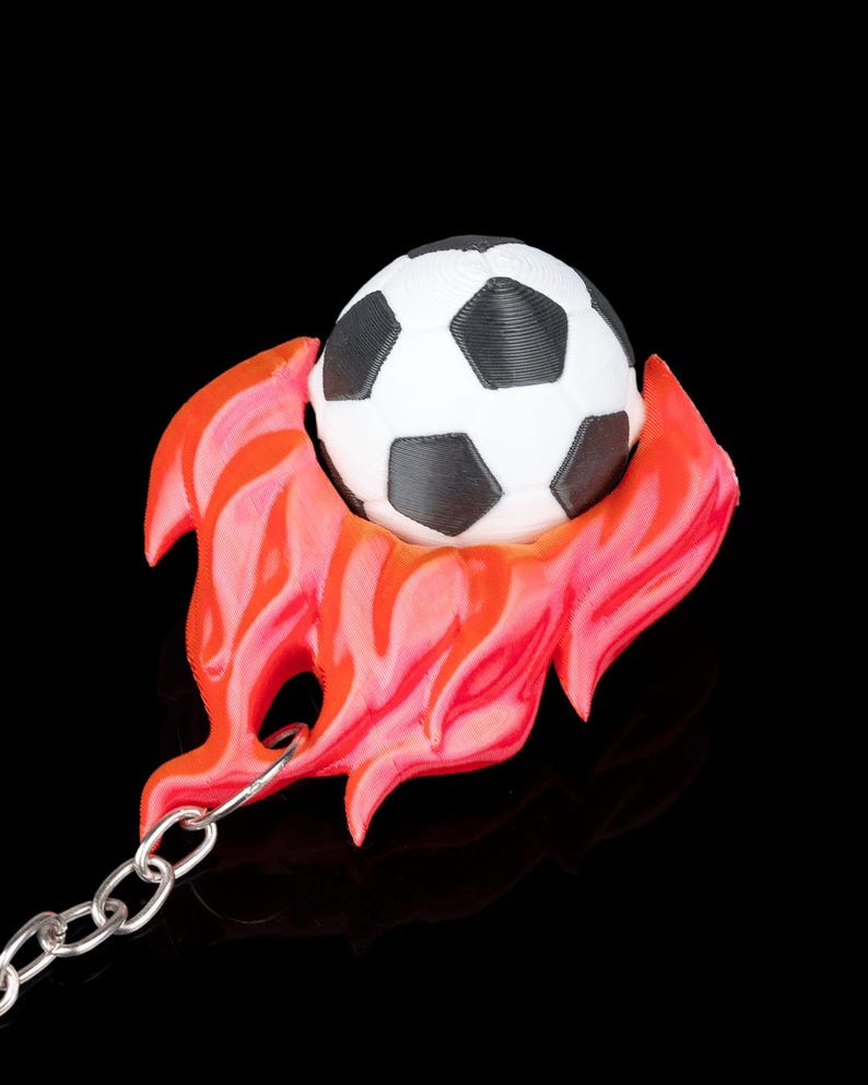 May include: A soccer ball keychain with a black and white soccer ball design, set within a red and orange flame-like shape. The keychain has a silver chain and a black background.