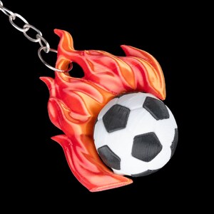 May include: A soccer ball keychain with a black and white soccer ball design, set against a backdrop of red and orange flames. The keychain is attached to a silver chain, creating a sporty accessory.