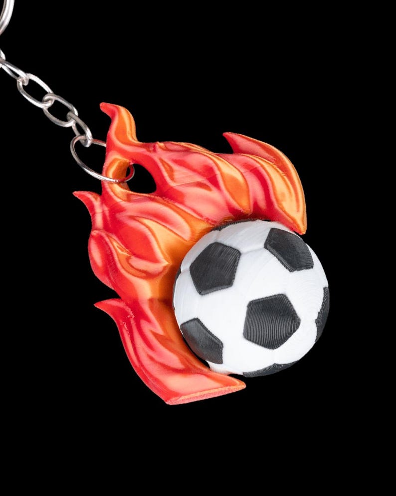 May include: A soccer ball keychain with a black and white soccer ball design, set against a backdrop of red and orange flames. The keychain is attached to a silver chain, creating a sporty accessory.