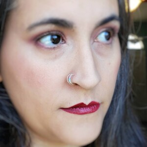 Puede incluir: Primer plano de un anillo de nariz plateado con forma de media luna, adornado con pequeñas piedras de color turquesa. La joya se lleva en la nariz de una persona, destacando el delicado diseño y el contraste de la plata sobre la piel.