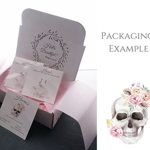 Puede incluir: Una caja blanca con el texto "Hello Beautiful!" y "Ready to rock?" impreso en la tapa interior. La caja contiene tarjetas de joyer&iacute;a y papel de seda rosa. Las tarjetas presentan dise&ntilde;os de joyas y tatuajes. Una ilustraci&oacute;n de calavera con flores est&aacute; a la derecha.