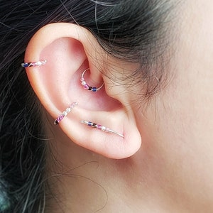 May include: Close-up of a person's ear with multiple ear piercings. The piercings are adorned with small, colorful beads and silver hoops.
