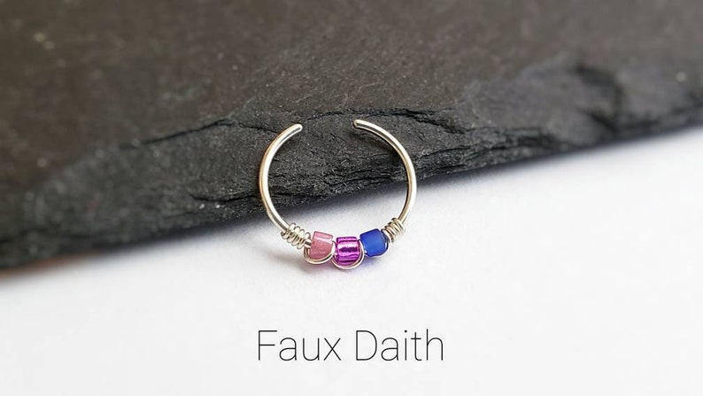 Puede incluir: Un pendiente falso daith de color plateado con tres peque&ntilde;as cuentas de colores. Las cuentas son rosas, moradas y azules. El pendiente tiene un dise&ntilde;o simple y circular. El texto "Faux Daith" est&aacute; debajo.