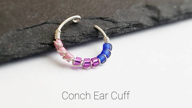 Puede incluir: Un pendiente de concha plateado con cuentas rosas, moradas y azules. El pendiente est&aacute; hecho de un alambre plateado que se enrolla alrededor de la oreja. El texto "Conch Ear Cuff" est&aacute; debajo.