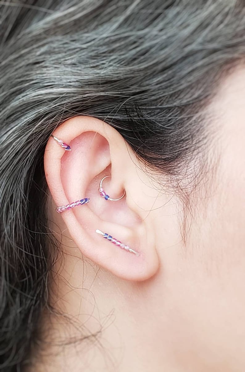 Puede incluir: Primer plano de una oreja adornada con m&uacute;ltiples piezas de joyer&iacute;a. Las perforaciones en la oreja presentan una variedad de pendientes, incluyendo un aro, una barra y una pieza curva, todos con acentos rosas y azules. La joyer&iacute;a se encuentra sobre un fondo de pelo gris.