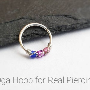 Puede incluir: Un pendiente de aro plateado con cuentas azules, rosas y moradas. El texto "20ga Hoop for Real Piercings" est&aacute; en la parte inferior de la imagen. El pendiente est&aacute; sobre una superficie gris oscuro.